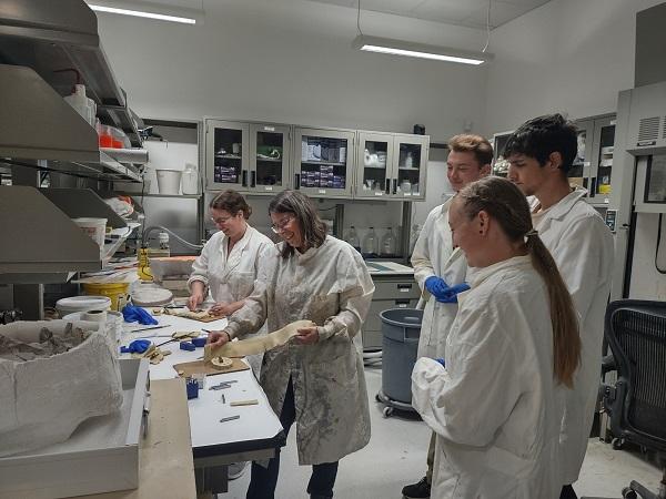 A woman (JODA fossil preparator) shows techniques for casting replicas of fossils to a group of three museum technicians who are looking on dressed in their lab coats. 