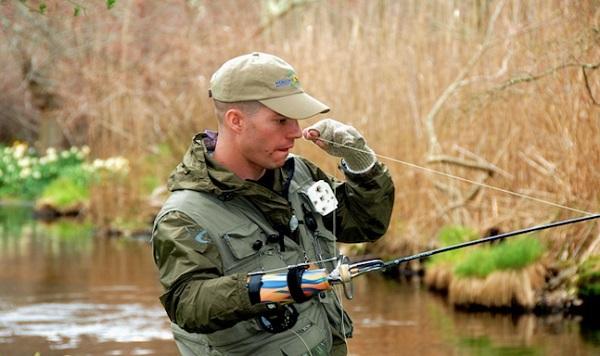 A military veteran is holding his fishing pole as he fishes in a stream. 