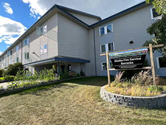 Photo of front of barracks gray building and entrance with an Alaska Fire Service sign in the summertime..