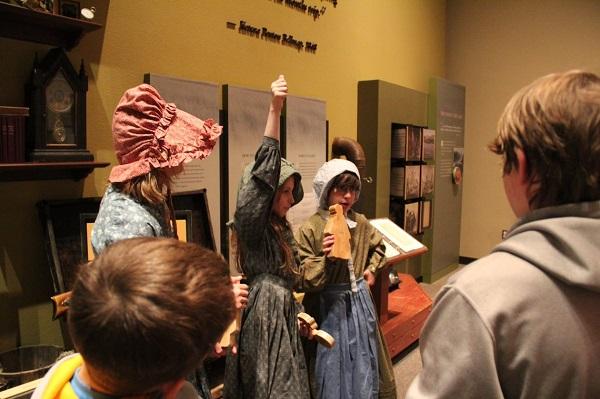 A girl dressed in period clothing with a dress and bonnet raises her arm as part of a presentation while other children look on.