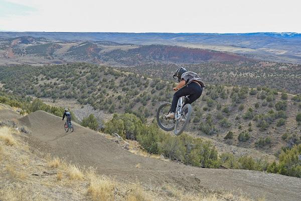 A rider hucks himself off a jump while another rider approaches a jump farther down the trail.