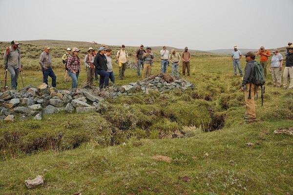 A group of people standing around and near some rocks and looking down at a dried-up stream covered in grass.