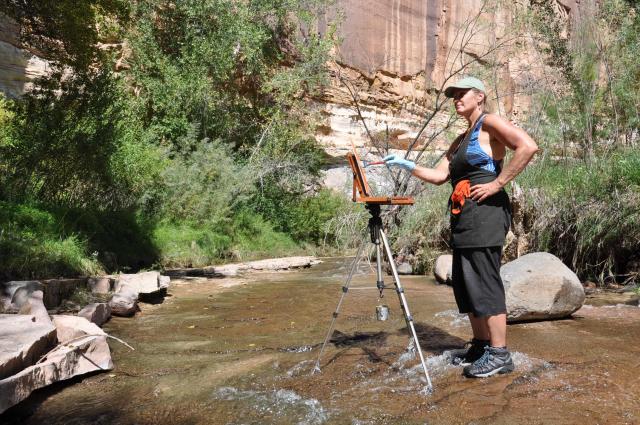Photo of female artist standing in a shallow, rocky stream and painting there on an easel