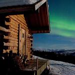 Green northern lights paint the sky behind a log cabin on a clear winter night.  Firewood is stacked on the front porch.  Moon shadows streak across the snow. 