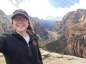 A girl with a black jacket, glasses and baseball cap, dark blond hair in a braid, stands at the top of a canyon. 