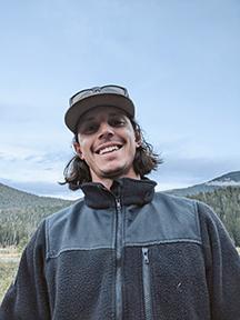 A man with blue jacket, baseball cap, and dark brown hair stands with forested hills behind him. 