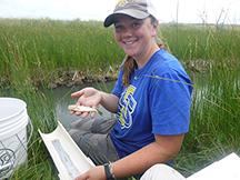 A young girl sits next to a creek, smiling at the camera, and holding a small minnow in her hand