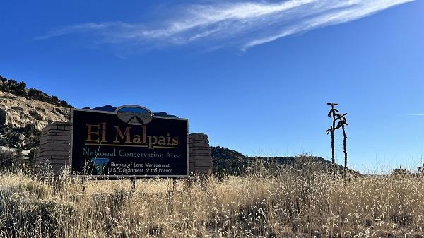 El Malpais National Conservation Area sign surrounded by dry brown grass under a blue sky. 