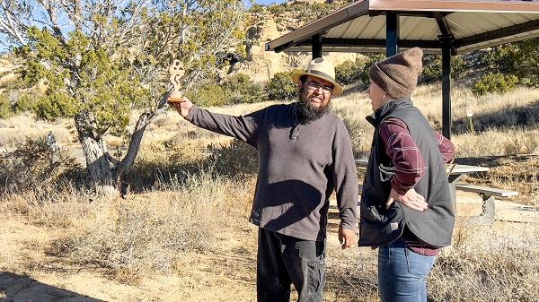 Walter Torres holds up a scaled down version of the statue and shows Kymm Gresset how the statue will frame the peak of Mount Taylor. There is a tree in the background and a patio with a picnic table. 