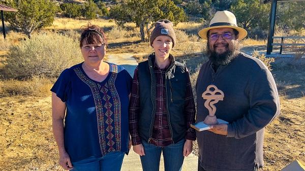 Left to right: Chief of Interpretation Lisa Dittman, El Malpais National Park; Kymm Gresset, Rio Puerco Field Office Manager; and Sculptor Walter Torres posing and smiling. Walter Torres is holding a scaled down version of the sculpture. There are some trees in the background.