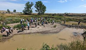 29 people stand on the bank of a muddy water source