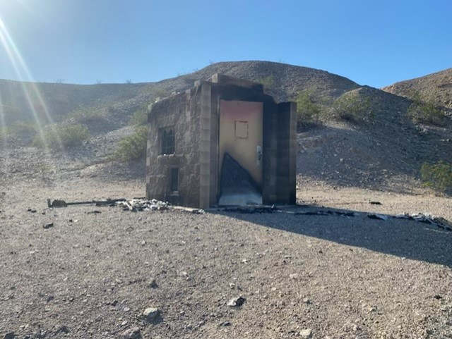 Burned vault toilet showing charred cinderblock walls and melted door