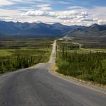 A gravel road runs up the mountain valley, paralleled by an oil pipeline. The grey road and shiny pipeline contrast with the greens of the spruce forest and wetland meadows. The road disappears in the row of mountains in the distance. The sky is marbled blue and white with high clouds. Underneath a few fluffy clouds build over the mountains. 