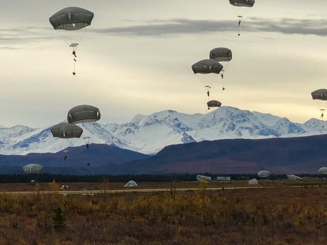 Paratroopers with open parachutes float through the sky and land on the grassy plain with snow-capped mountains in the background.