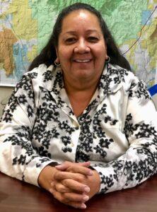 A woman smiles at the camera while sitting at a desk with her fingers interlaced. 