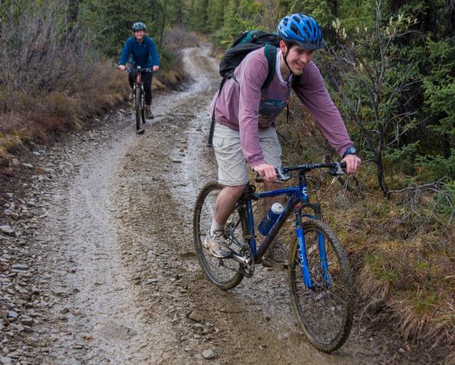 Two mountain bike riders coasting downhill on a two-track, multi-use trail through the woods. 