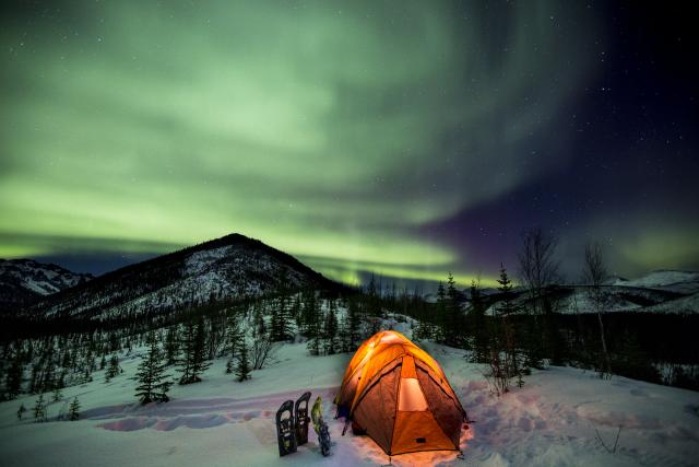 A winter campout under the northern lights. A yellow tent glows with light from within while the green lights fill the night sky.