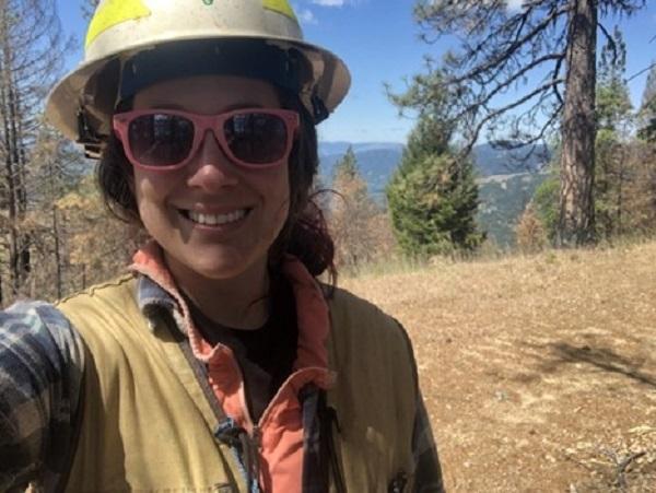 A woman stands in a forest with a hard hat on. 