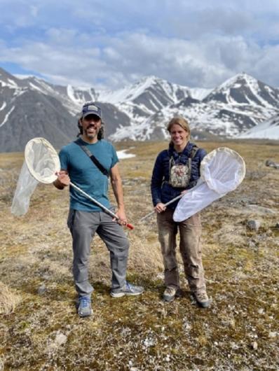 A man and a woman stand with bee nets with mountains in the background. 