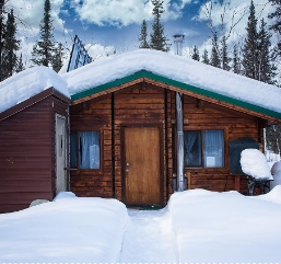 Coldfoot cedar cabin during the winter. Snow is piled on the roof and around the cabin. A narrow trail leads to the front door.