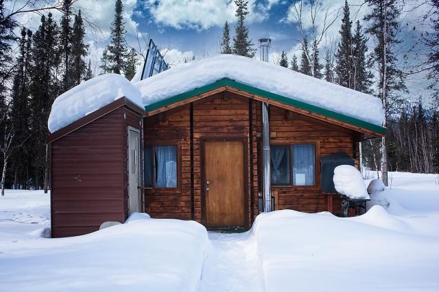 Coldfoot cedar cabin during the winter. Snow is piled on the roof and around the cabin. A narrow trail leads to the front door.