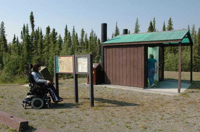 One person in a wheel chair reads a sign while another enters a restroom. 