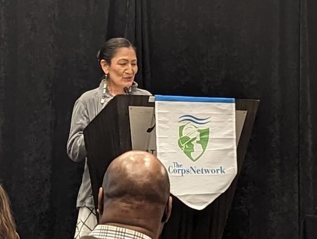 Secretary of the Interior, Deb Haaland, standing at a lectern with a small banner hanging from it which says the Corps Network