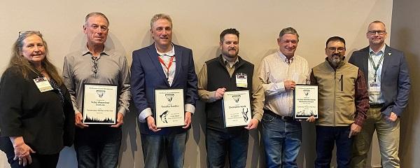 A woman on the left and then 6 men are lined up holding their awards. 