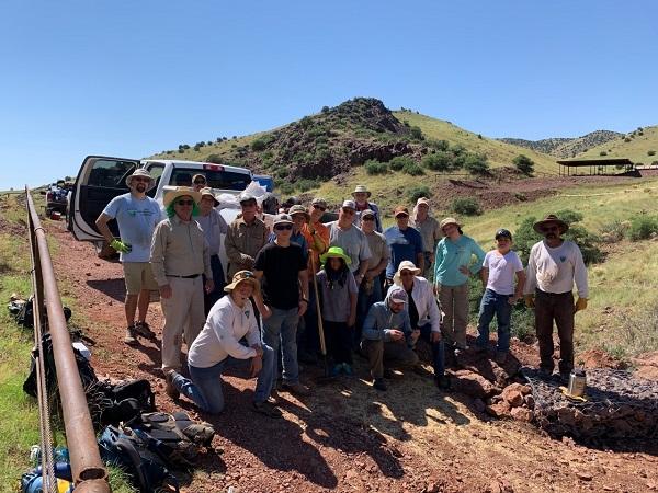 A large group of Socorro Field Office staff and volunteers pose on a dirt road with a green hill in the background. 