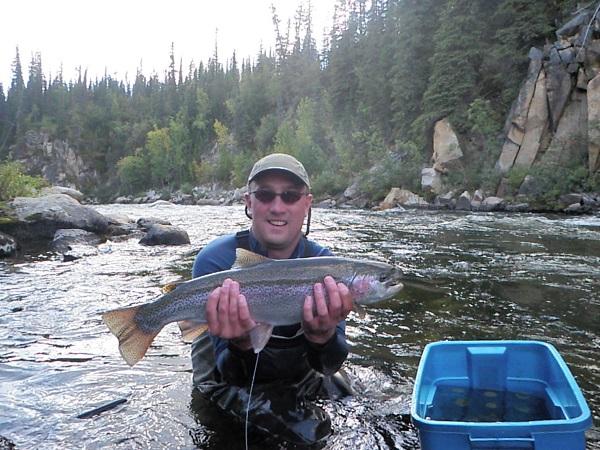 A man stands in a creek holding a fish and smiling. Next to him is a blue bucket.