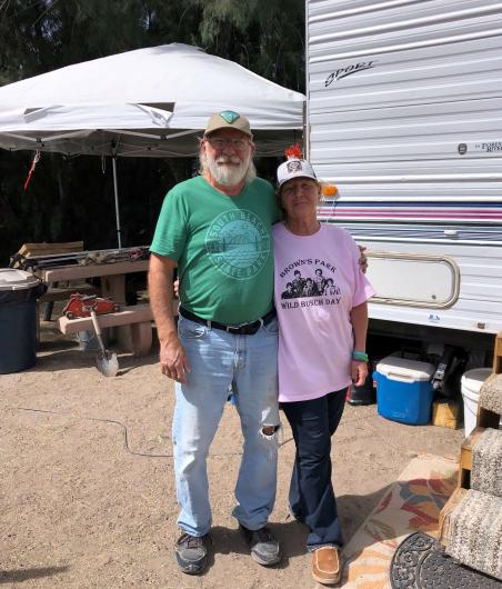 Two people wearing baseball caps stand next to a camper and in front of a picnic table