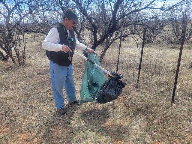 Man wearing a cap and vest with the BLM logo using a trash picker to remove a garbage bag full of trash from the ground