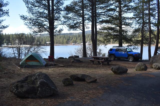 campsite with tent, fire pit, and picnic table, surrounded by fir trees, with body of water in the background