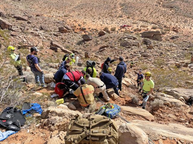 Group of people in a rocky desert area. Some are wearing bright yellow shirts and yellow helmets and several ropes are visible..