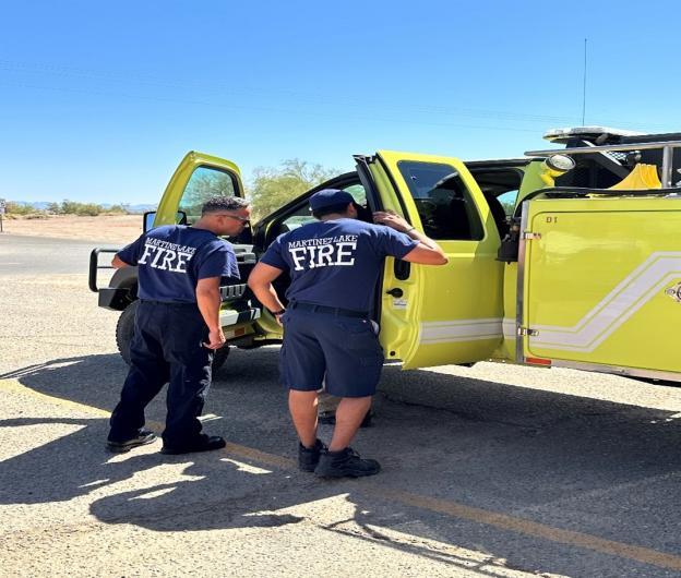 Two people wearing shirts that say Martinez Lake Fire on the back are looking inside a fire truck engine that has its side doors open.