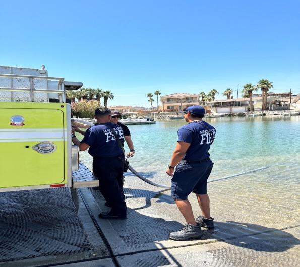 Three fire personnel stand next to a yellow fire engine. The end of the engine's water hose is submerged in a lake.