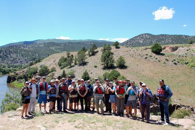 Group image of BLM employees and NW RAC members, with the Colorado River and rolling hills in the background. Everyone is wearing a lifejacket and hat. 