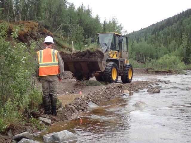 A person in an orange reflective vest and white hard hat directs heavy equipment operator along stream bank.