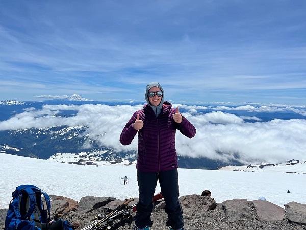 A woman gives the thumbs up sign on top of a snowy mountain. 