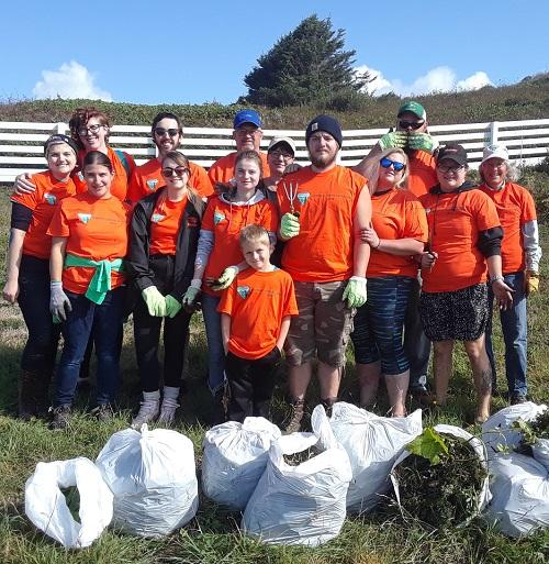 group of volunteers of all ages, all wearing orange shirts, posing in front of bags of weeds that they collected 