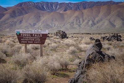 A sign reading "Fossil Falls Campground" in the high desert under snow capped peaks