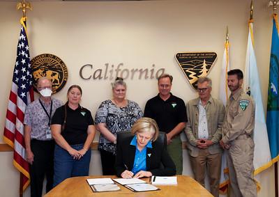 A woman sits at a desk and signs a document while six adults stand behind her looking on. 