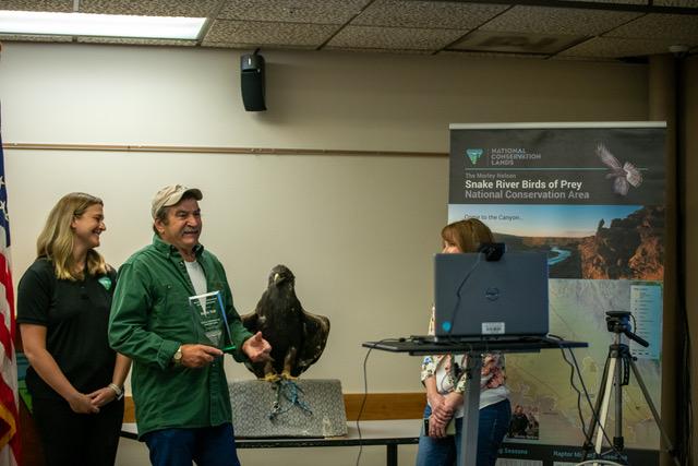 A man holds a an award plaque in the other. A raptor stands on a box next to him. To his left a woman is smiling. To his right, another woman stands behind a laptop screen. A Snake River Birds of Prey sign is in the background. 