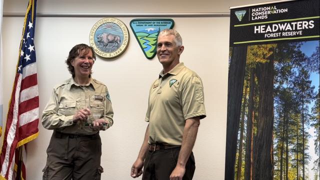 A woman holds a plaque smiling. To her left is a man smiling and the Headwaters Reserve sign. 