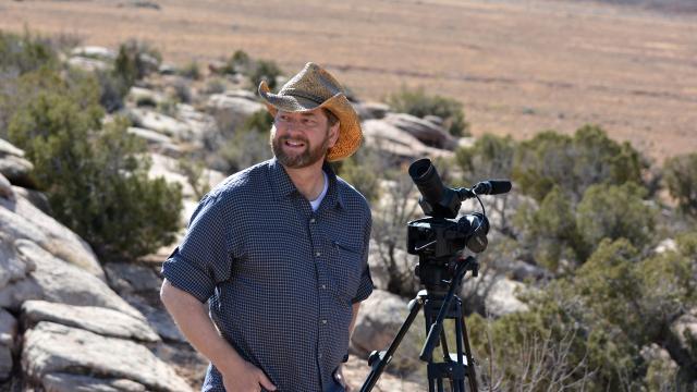 A photographer in a dark blue button-down shirt and cowboy hat grins next to a camera pointed toward the sky.