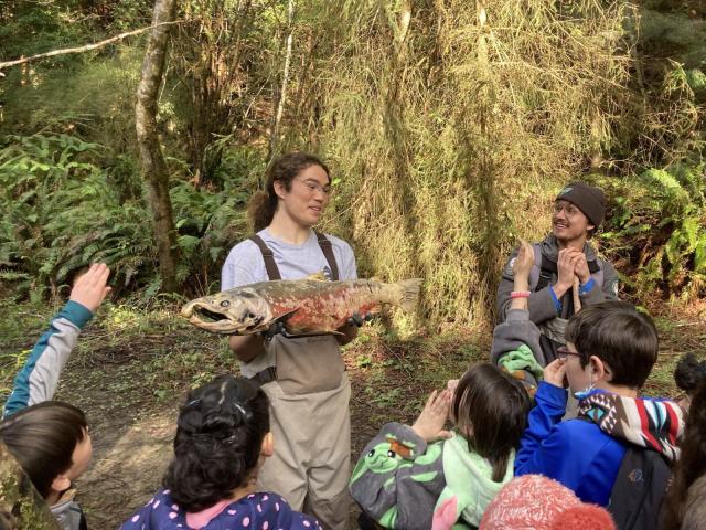 A volunteer holds a large salmon in front of a group of children.