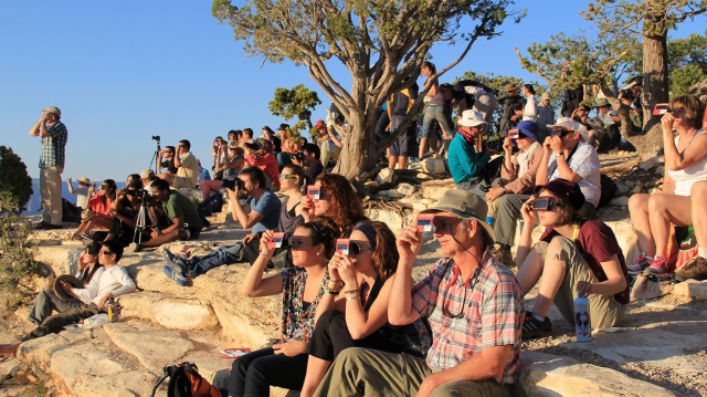 a crowd of people sitting on sandstone amid juniper trees hold paper glasses up to their faces.