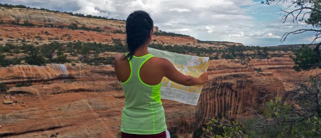 A woman in a yellow tank top looks out over a sandstone canyon holding a map in her hands.
