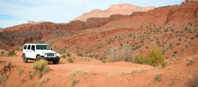 A white jeep climbs an orange dirt road with massive sandstone cliffs in the background.