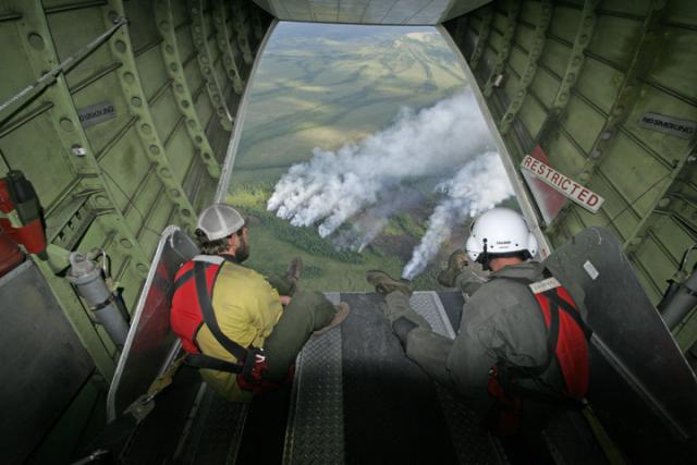 Two people sit on the end of an open back end of an airplane watching smoke billow from a forest fire. 